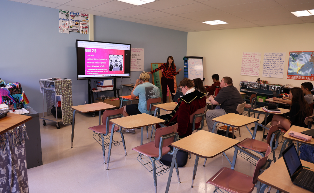 teacher standing next to a smart board in front of a classroom of students who are sitting at desks